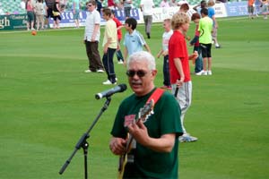 man singing into microphone with guitar on cricket outfield